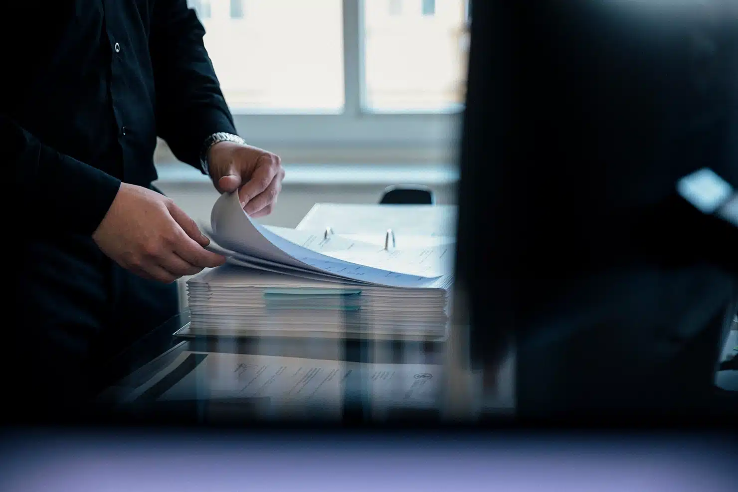Person in dunkler Kleidung blättert in ringgebundener Akte auf dem Schreibtisch; Fenster im Hintergrund