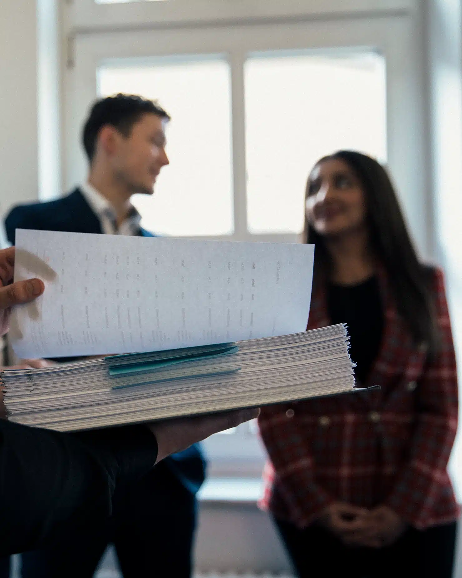 Person hält dicken Aktenstapel; im Hintergrund zwei unscharfe Personen in hellem Büro vor Fenstern.
